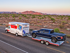 A U-Haul moving truck driving on the highway towing a truck on a U-Haul utility trailer