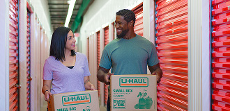 A couple standing in a U-Haul storage facility each holding a packed box while smiling at each other.